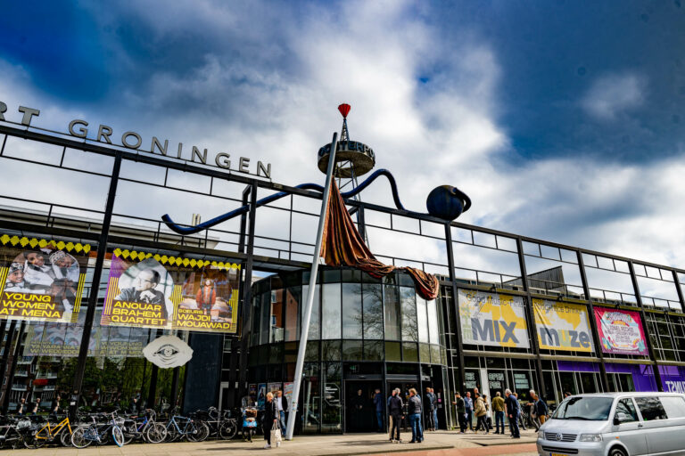 Een modern gebouw met een opvallende kunstinstallatie op het dak en mensen die naar de ingang lopen.