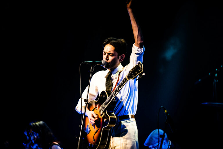 Een muzikant met een gitaar zingt en steekt zijn hand omhoog op het podium.