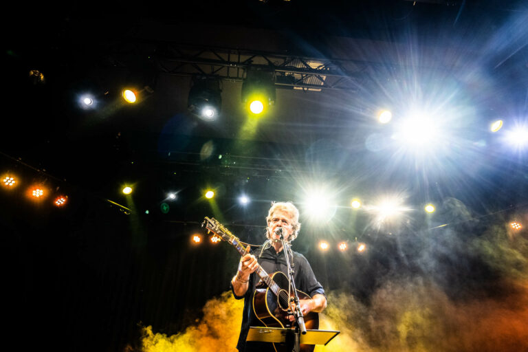 Een muzikant met een gitaar staat op het podium omringd door fel licht en rook.