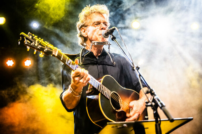 Een muzikant speelt gitaar en zingt op een podium, omringd door kleurrijke verlichting.
