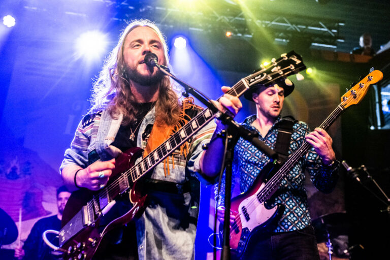 Twee muzikanten staan op het podium en spelen gitaar tijdens een live optreden met kleurrijke verlichting op de achtergrond.