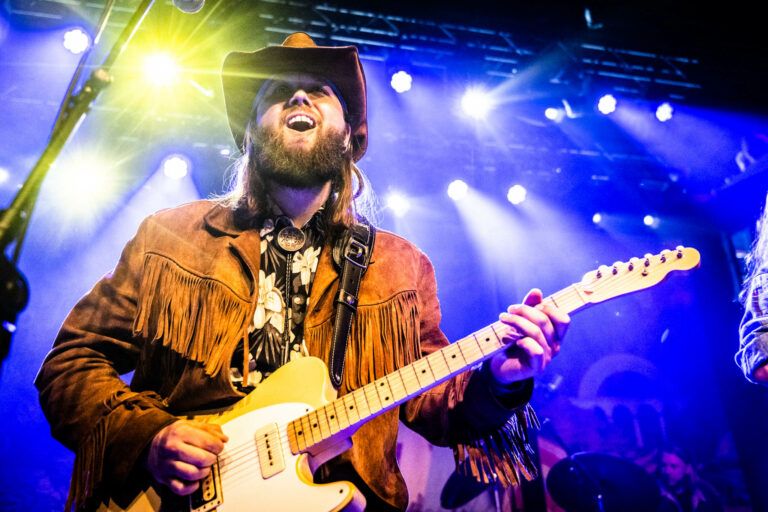 Een muzikant in cowboyhatt en een fringed jas speelt op een elektrische gitaar op een kleurrijk verlichte podium.
