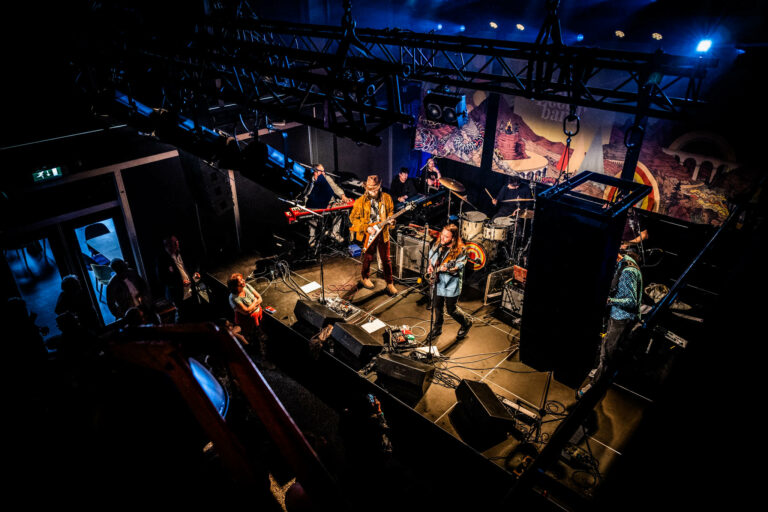 Een live band speelt op een podium met gekleurde verlichting en een publiek in de schaduw.