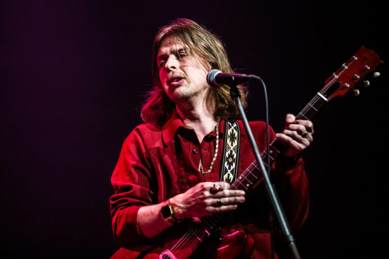 Een muzikant met lang haar in een rood shirt speelt gitaar en zingt in een microfoon.