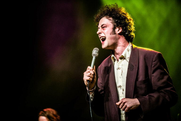 Een zanger met krullend haar staat op een podium terwijl hij in een microfoon zingt, met een kleurrijke achtergrond.