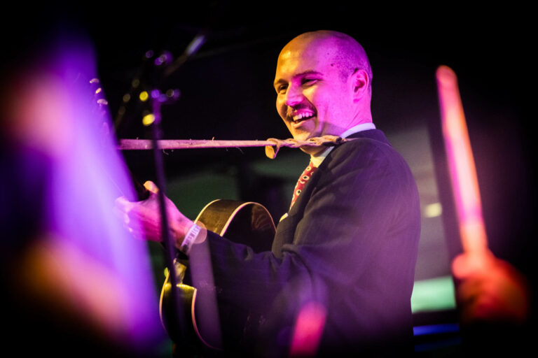 Een muzikant die lachend een gitaar speelt op een kleurrijk verlichte scène.