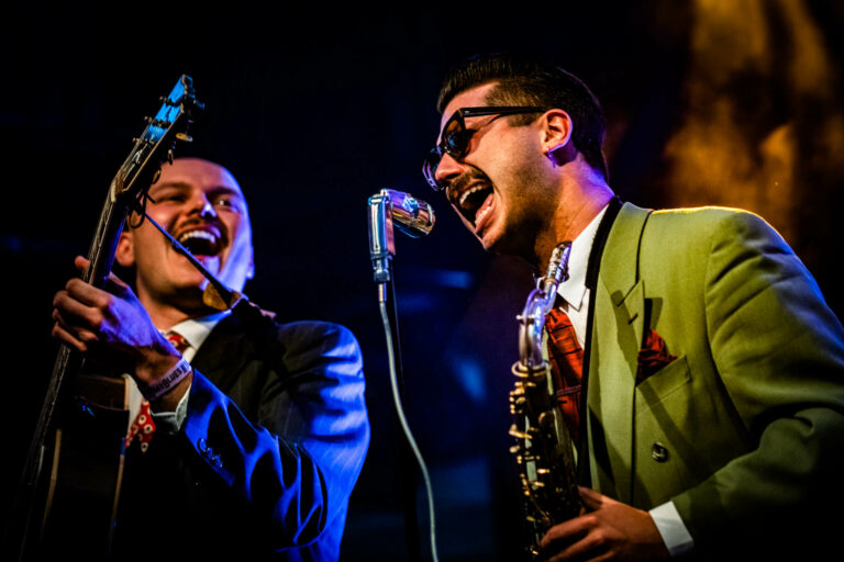 Twee muzikanten met een sax en microfoon optreden op het podium, gekleed in stijlvolle kostuums.