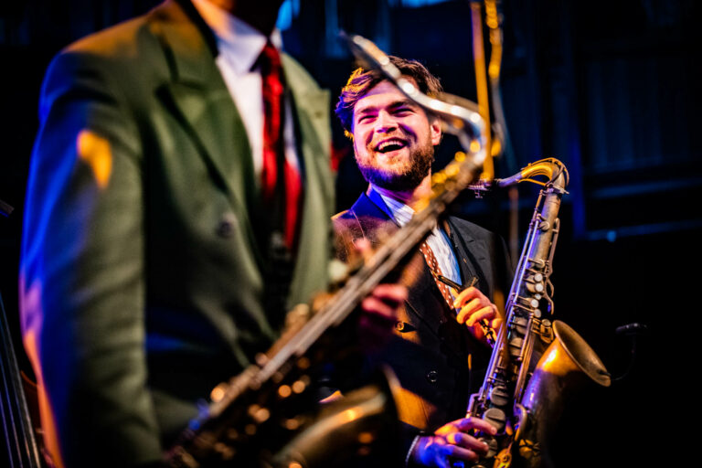 Twee saxofonisten spelen op een podium, met de focus op een glimlachende muzikant op de voorgrond.