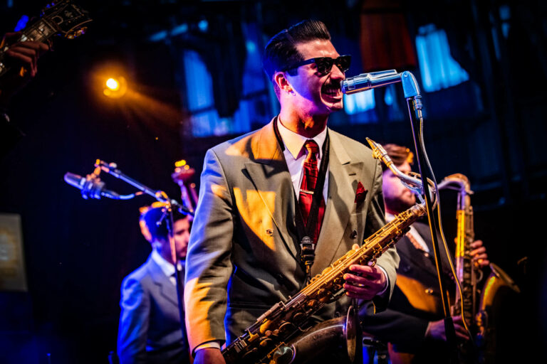 Een muzikant in een vintage kostuum speelt saxofon en zingt in een microfoon tijdens een optreden op het podium.