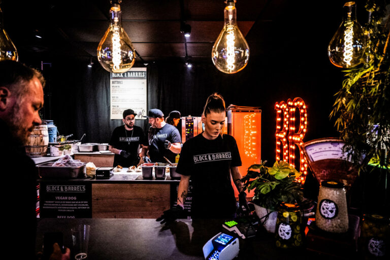 Een medewerker staat achter de kassa in een sfeervolle café-omgeving met sfeervolle verlichting en planten.
