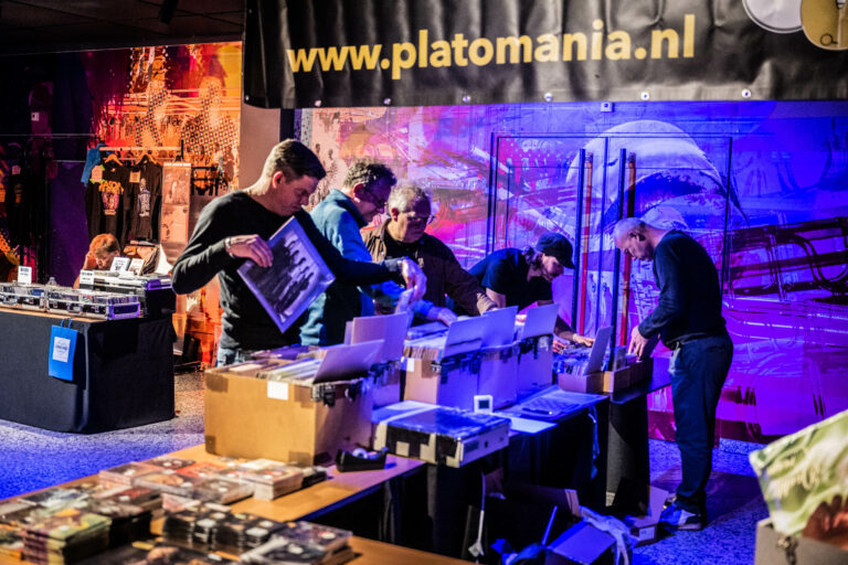 Een groep mensen aan een tafel bezig met het sorteren en verkopen van platen, met een kleurrijke achtergrond en een banner van Platomania.