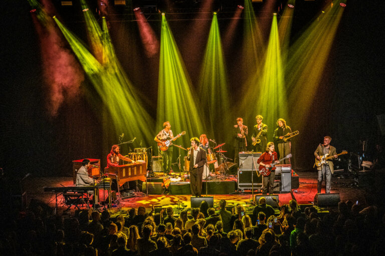 Een band speelt live op een podium, omringd door kleurige lichten en een menigte toeschouwers.