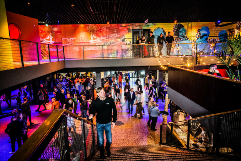 Een drukke binnenruimte met mensen die de trap afkomen, kleurrijke verlichting en een levendige sfeer.