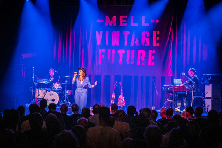 Een zangeres voert op een podium met een band, voor een enthousiast publiek, verlicht door kleurrijke lichten en een opvallend logo.