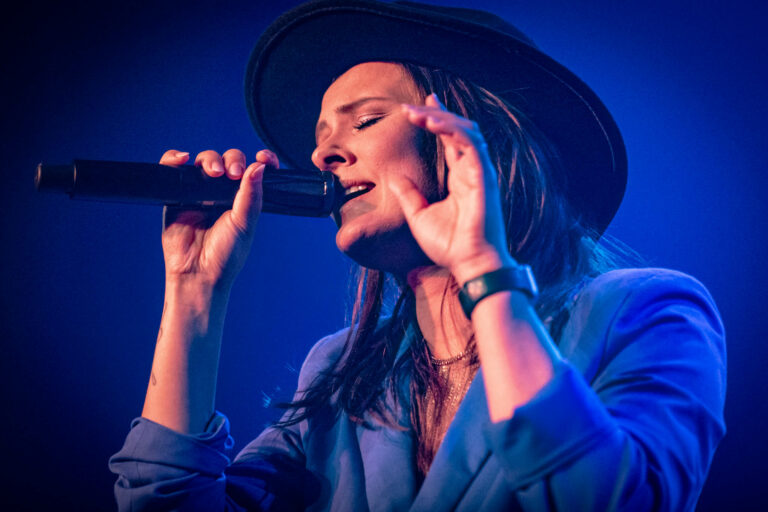Een vrouw in een blauwe blazer zingt met een microfoon, terwijl ze emotioneel met haar handen gebaren maakt op een podium.