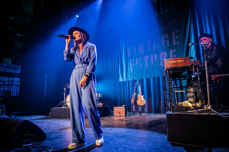 Een vrouwelijke zangeres in een blauwe jumpsuit staat op een podium onder blauwe verlichting.