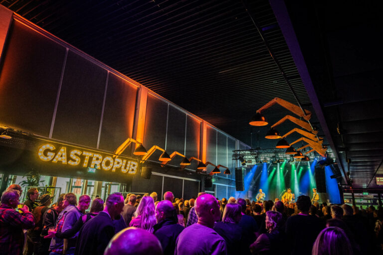 Een drukke evenementenlocatie met mensen die genieten van een optreden, onder kleurrijke verlichting en een sign van