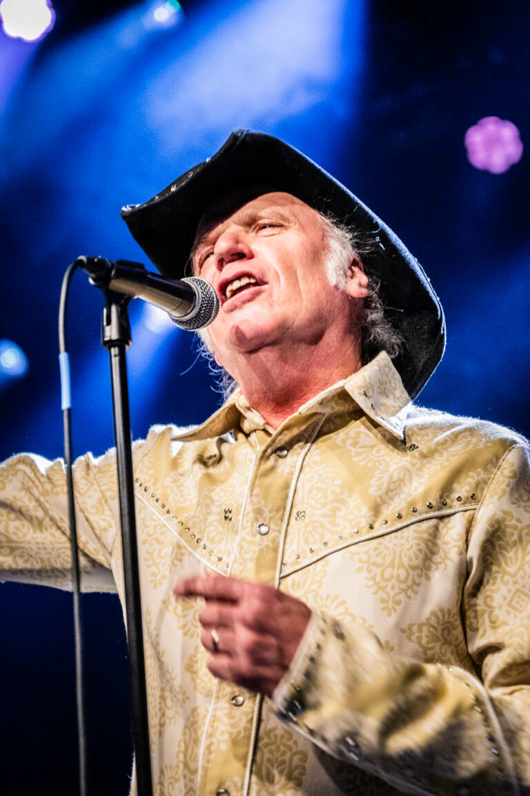 Een man met een cowboyhoed zingt enthousiast in een microfoon op een podium met blauwe verlichting.