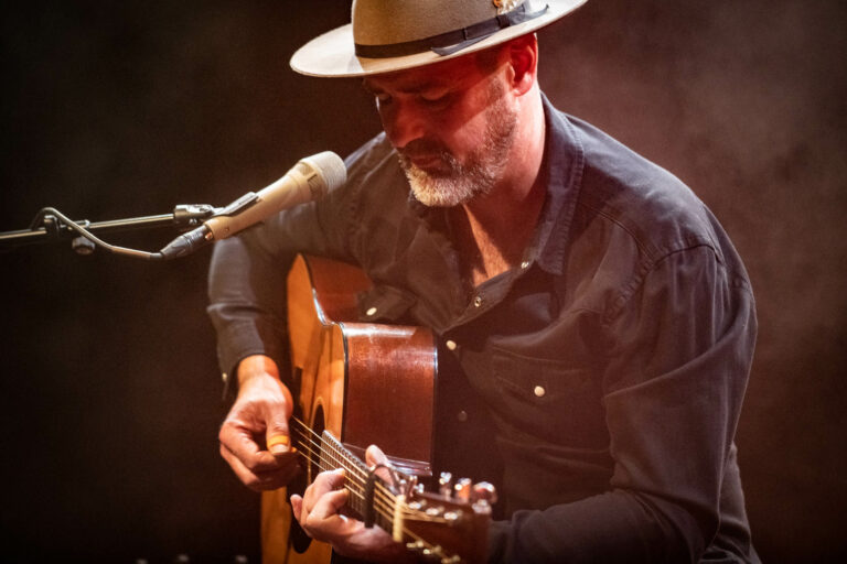 Een man met een hoed speelt gitaar en zingt in een sfeervolle omgeving.