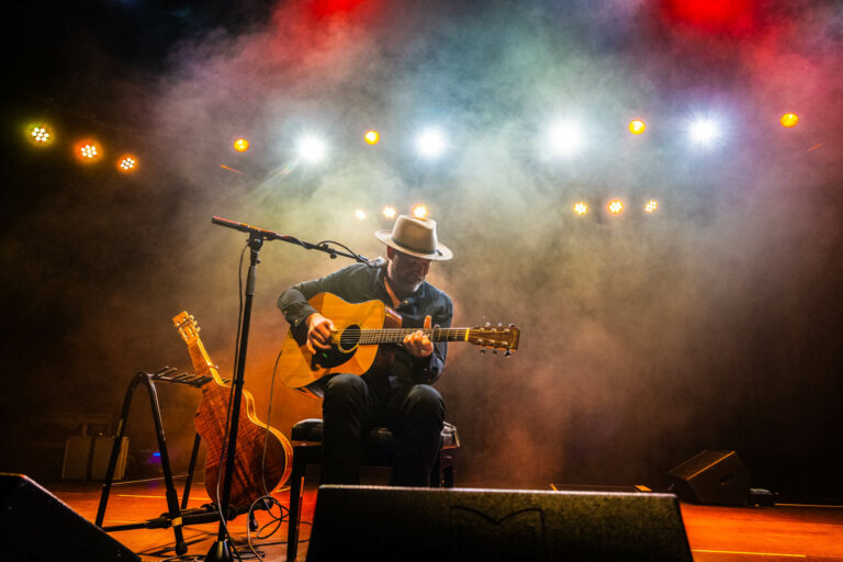 Een muzikant speelt gitaar op een podium, omringd door rook en kleurrijk licht.