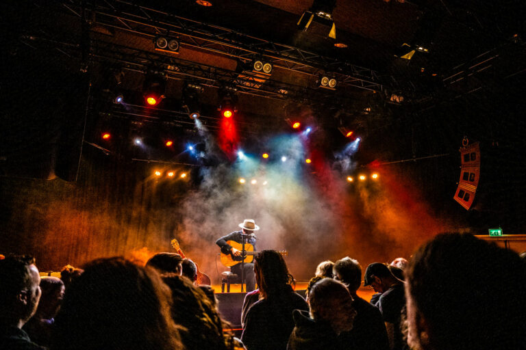 Een muzikant speelt gitaar op een podium met kleurrijke verlichting en een publiek in de schaduw.