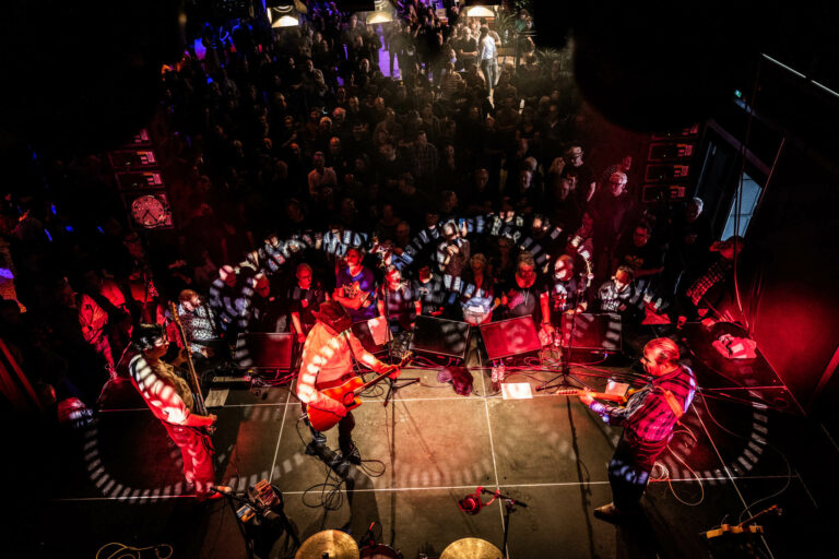 Een bovenaanzicht van een live muziekoptreden met een volle menigte en kleurrijke lichten.