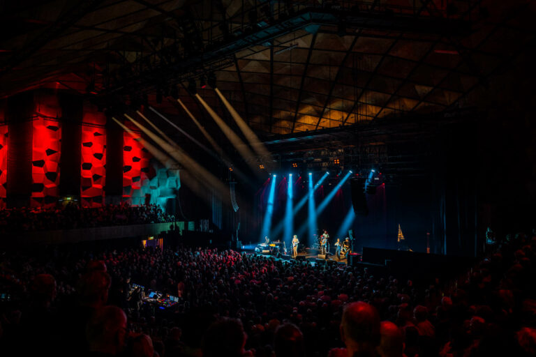 Een druk concert met een band op het podium, omringd door kleurrijke lichten en een uitgelaten menigte.