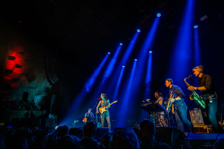 Een band speelt live op het podium met blauwe verlichting en een publiek ervoor.