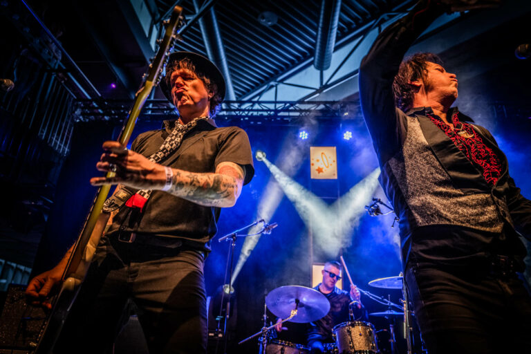 Twee muzikanten voeren op een podium met felgekleurde lichten en een drumstel op de achtergrond.