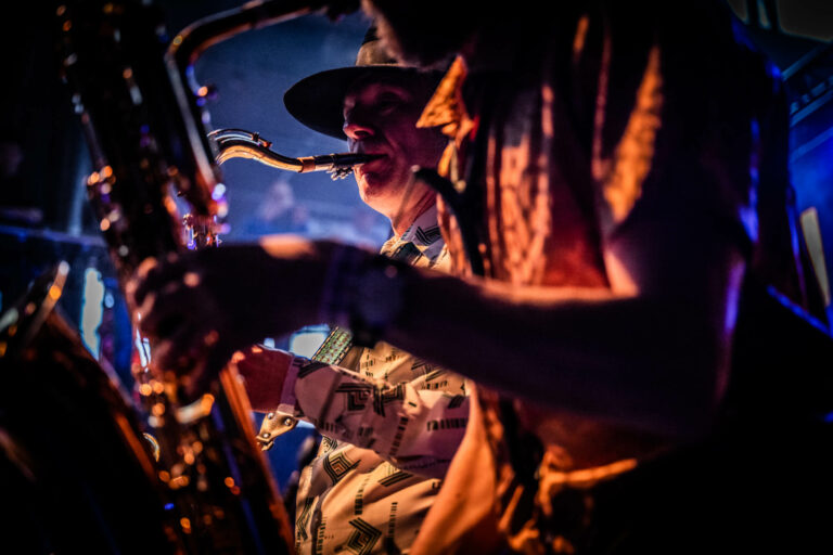 Twee muzikanten spelen samen op een podium, waarbij een saxofonist op de voorgrond zichtbaar is.