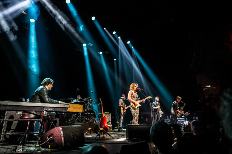 Een band speelt live op een podium, omringd door kleurrijke lichtstralen en muzikanten.