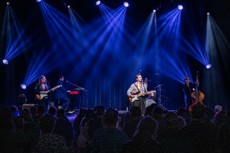 Een band speelt live op een podium met kleurrijke lichtstralen en een publiek dat geniet van de muziek.
