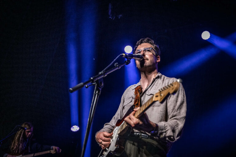 Een muzikant speelt gitaar en zingt op een podium, omringd door blauwe lichten.