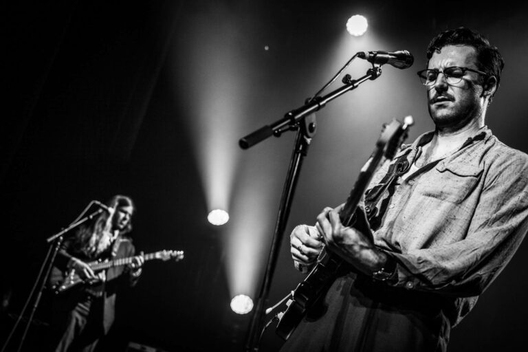 Twee muzikanten optreden op een podium, met focus op de gitaar en mic in een zwart-wit setting.