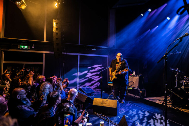 Een muzikant speelt gitaar op een podium terwijl het publiek enthousiast toekijkt.