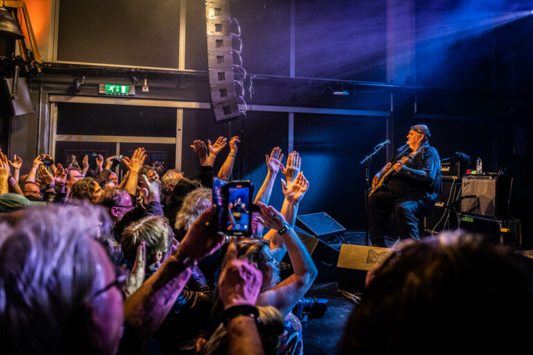 Een muzikant speelt op het podium voor een enthousiast publiek met opgestoken handen en telefoons die foto's maken.