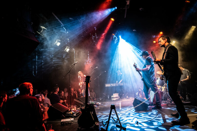 Een band speelt live voor een enthousiast publiek onder kleurrijke lichtstralen en mist.