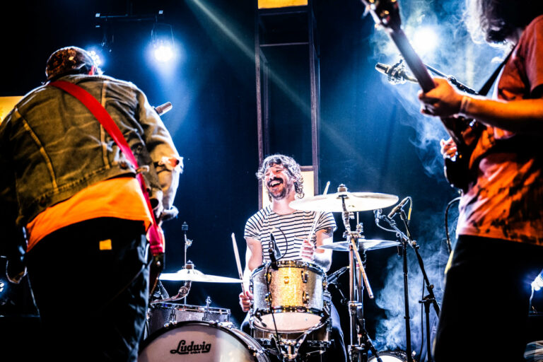 Een energieke band met een drummer en twee gitaristen die optreden op een verlichte podium.