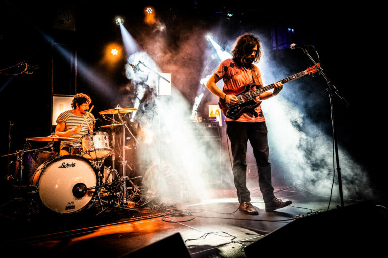 Een live band speelt op een podium met kleurrijke lichten en rook, terwijl de gitarist en drummer in het spotlight staan.