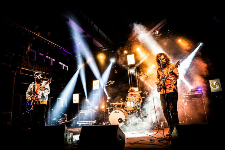Een band speelt live op het podium met kleurrijke lichtstralen en rook effect.