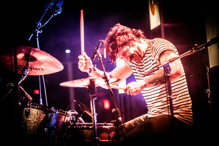 Drummer in een gestreept shirt speelt enthousiast op een drumstel tijdens een optreden.