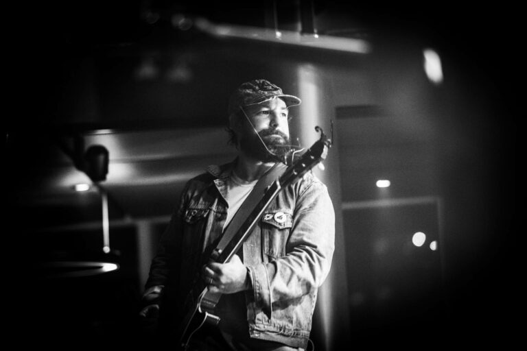 Zwart-wit afbeelding van een muzikant met een baard, die een gitaar vasthoudt tijdens een optreden.