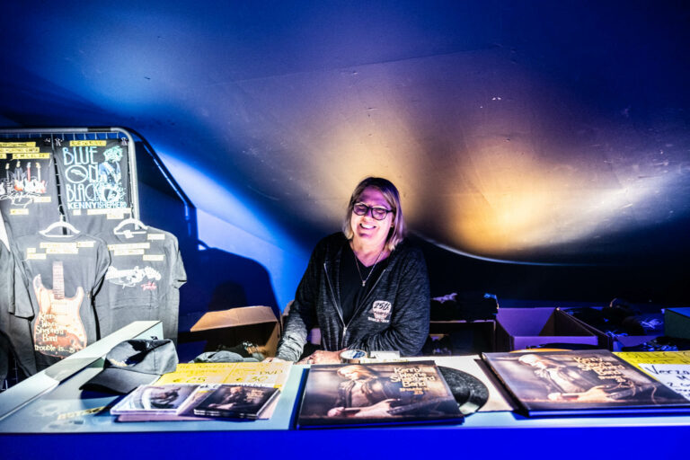 Een persoon staat achter een verkoopstand met merchandise en platen.