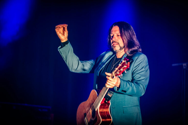 Een muzikant in een blauw kostuum speelt gitaar en steekt zijn hand in de lucht tijdens een optreden.