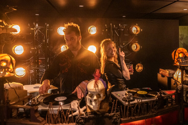 Een DJ-duo staat achter draaitafels, omringd door sfeervolle verlichting en een opvallende schedel.