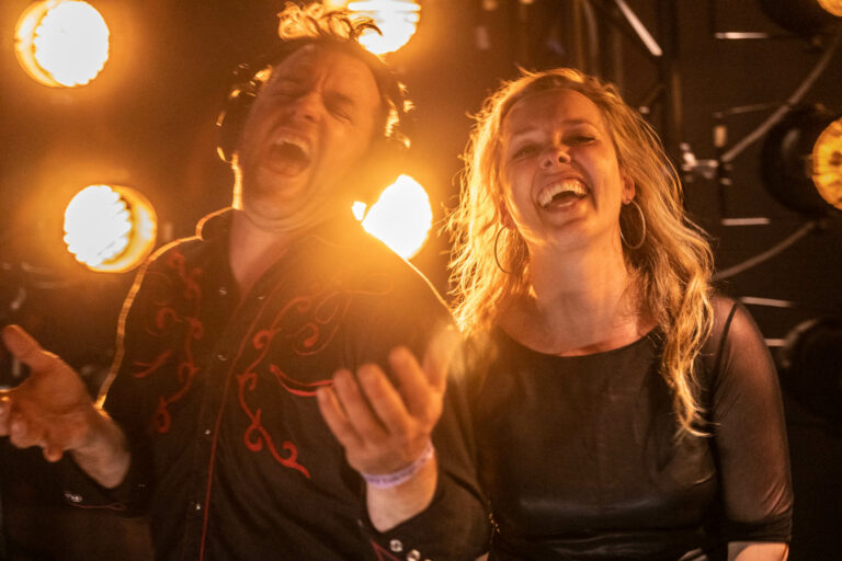 Twee lachende performers op een podium, omringd door warme lichtstralen.