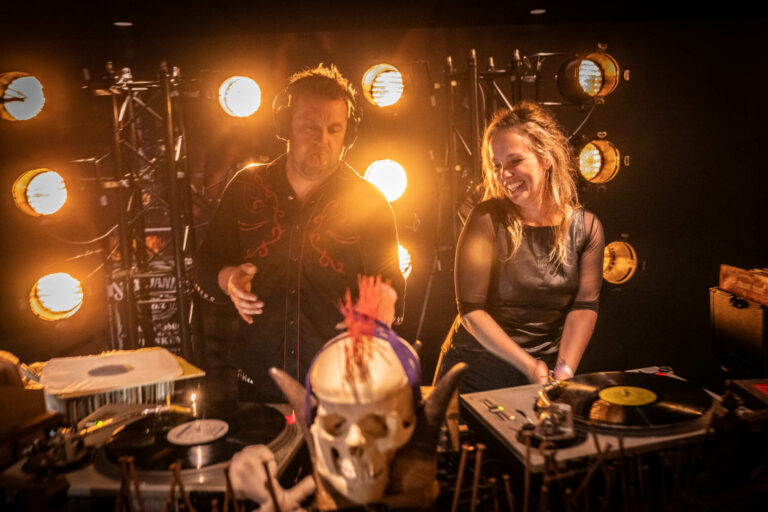 Een DJ-duo staat aan een draaitafel met muziekapparatuur en een decoratieve schedel, verlicht door warme lichten.