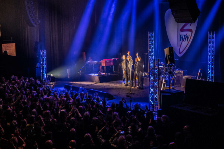 Een band speelt op het podium voor een enthousiast publiek onder blauwe lichtstralen.
