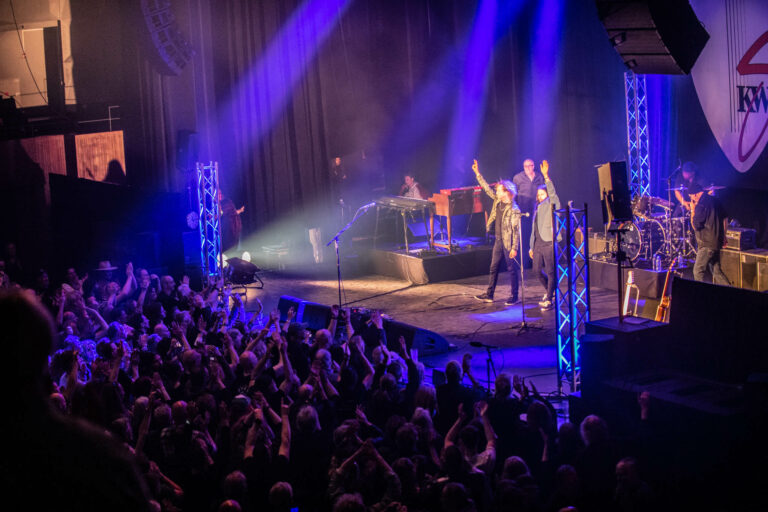 Een live muziekoptreden met een band op het podium, omringd door een enthousiaste menigte en kleurrijke lichten.