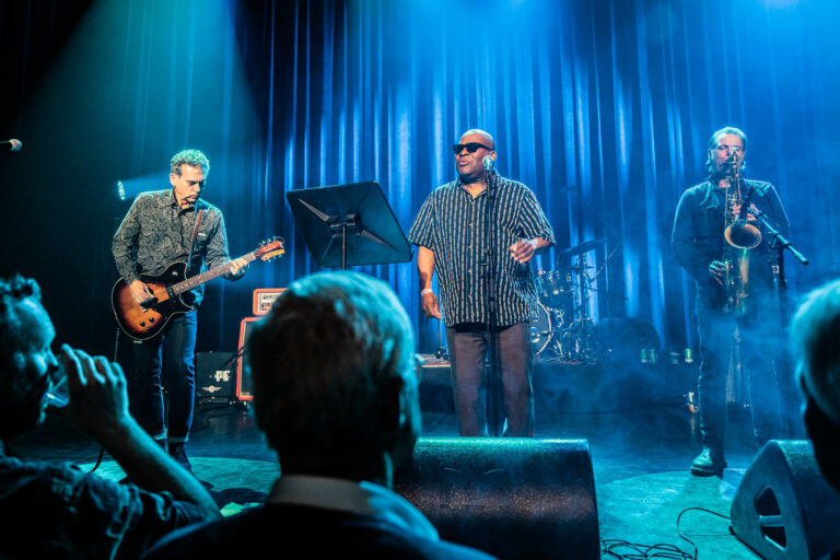 Een bandoptreden met een zanger en een gitarist in een met blauwe lichten verlichte zaal.
